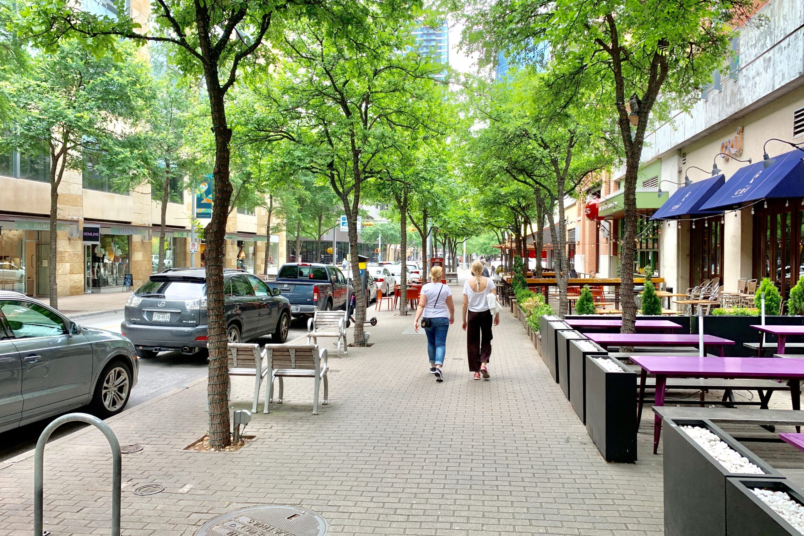 Two women in white shirts walking down a wide sidewalk with outdoor shops and purple benches
