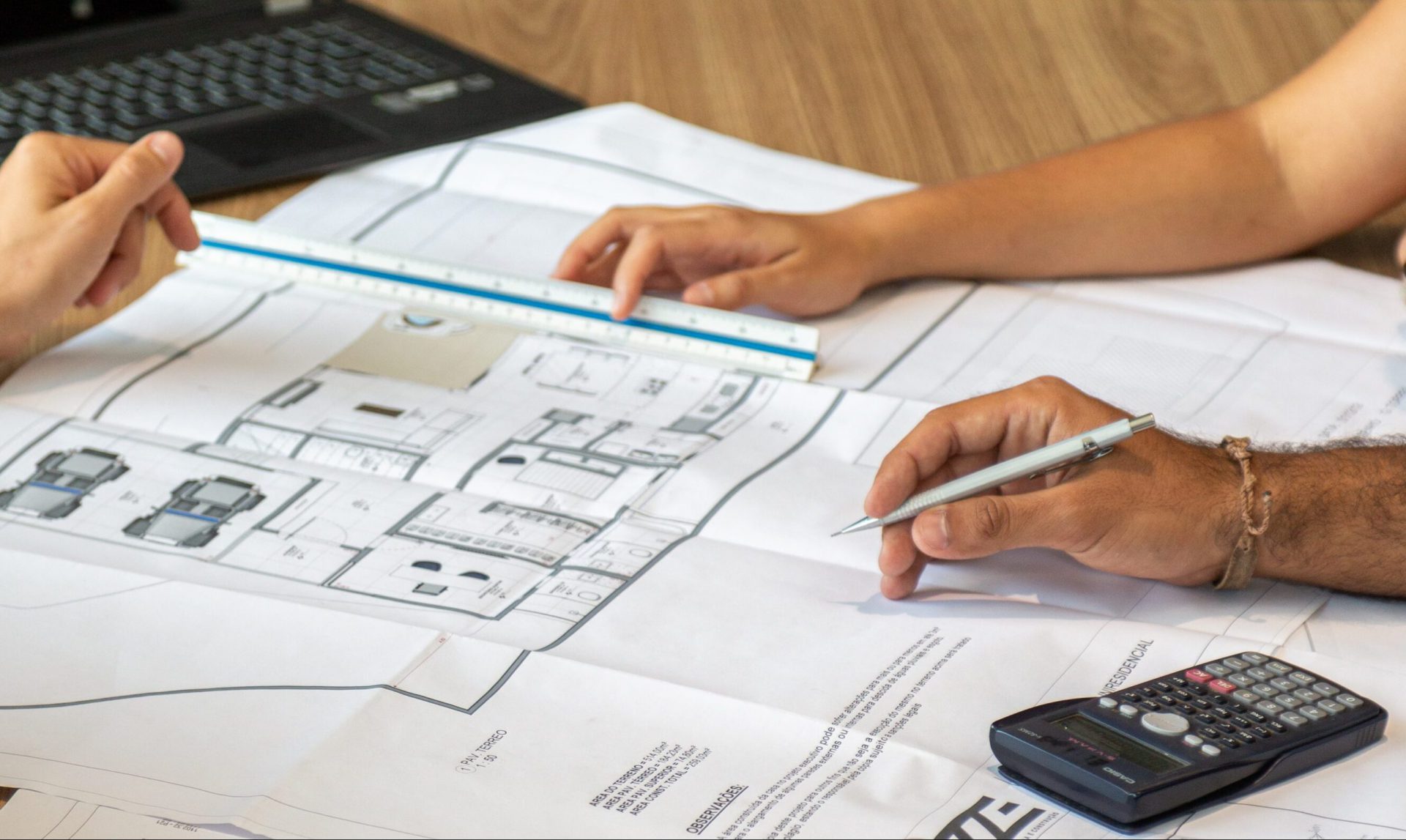 Floor plans on a wooden table with hands resting on top of it and a laptop in the background