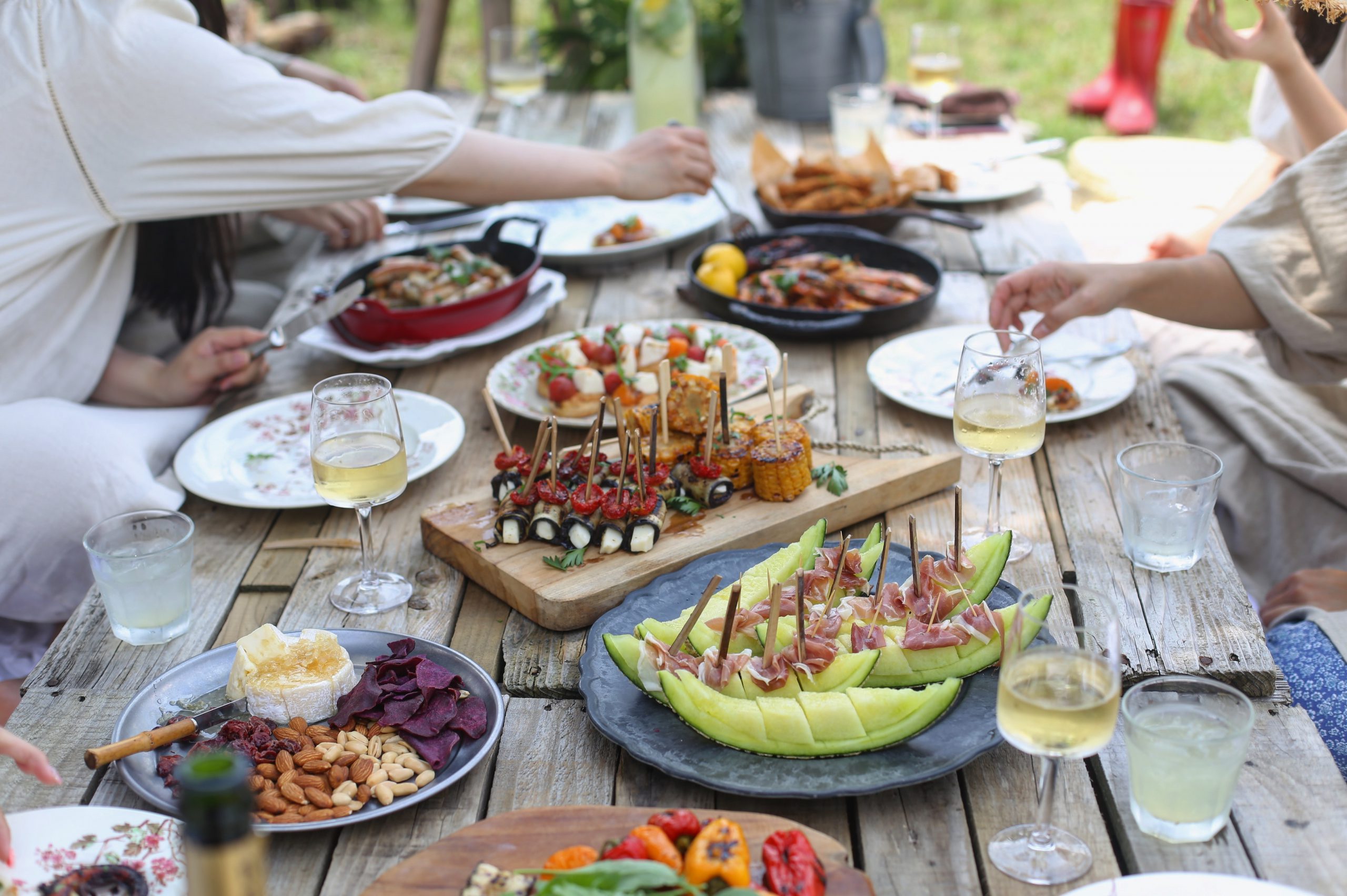 Variety of colorful foods on a wooden table with wine glasses and multiple hands reaching across
