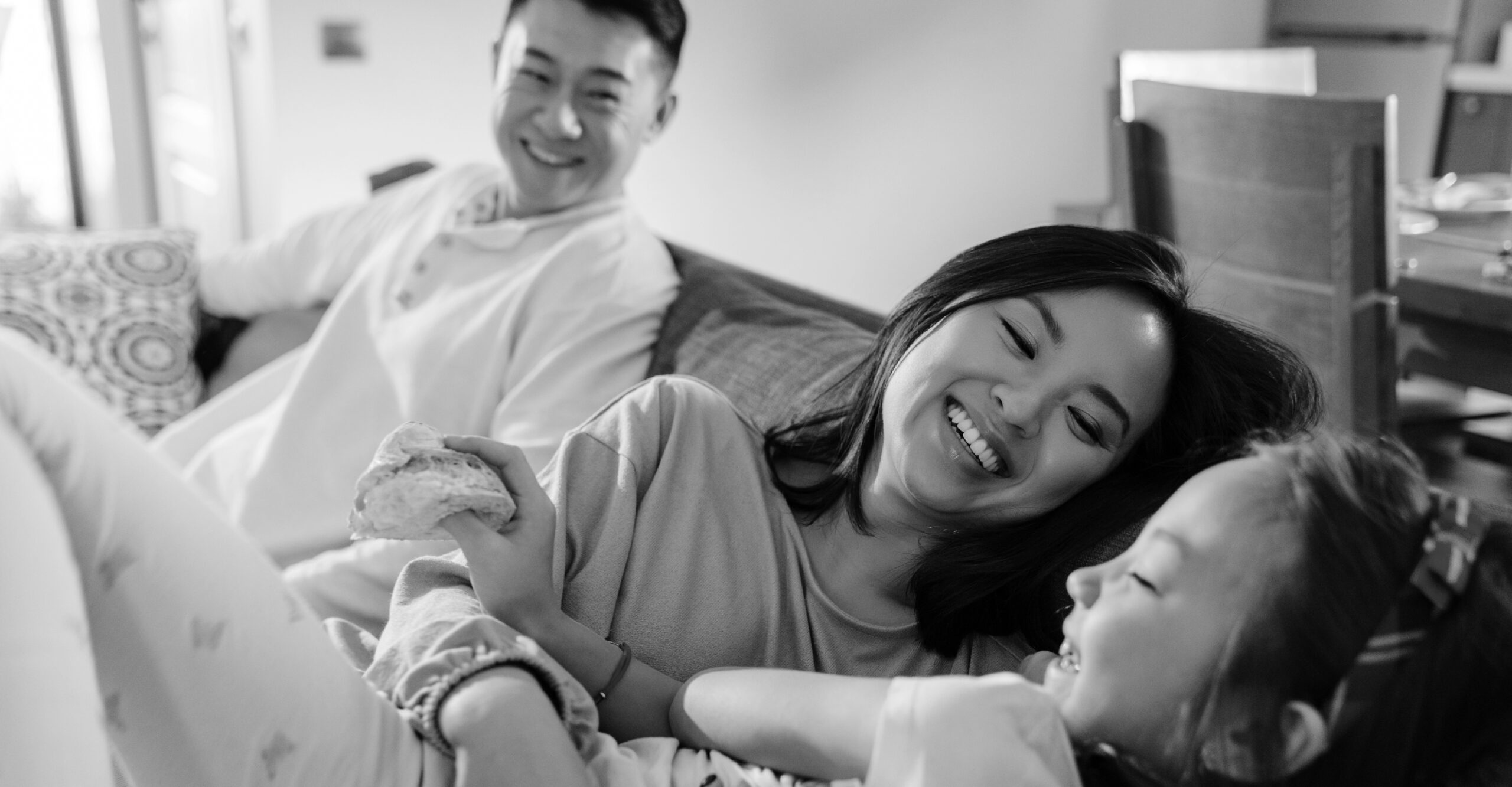Close up of a mom holding a small girl while they both laugh and a dad looking at them in the background